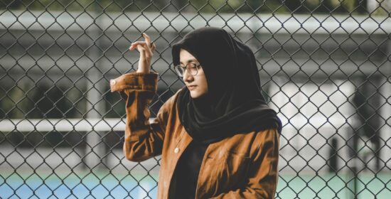 selective focus photography of woman wearing black hijab scarf and brown coat leaning on metal chain-link fence during daytime